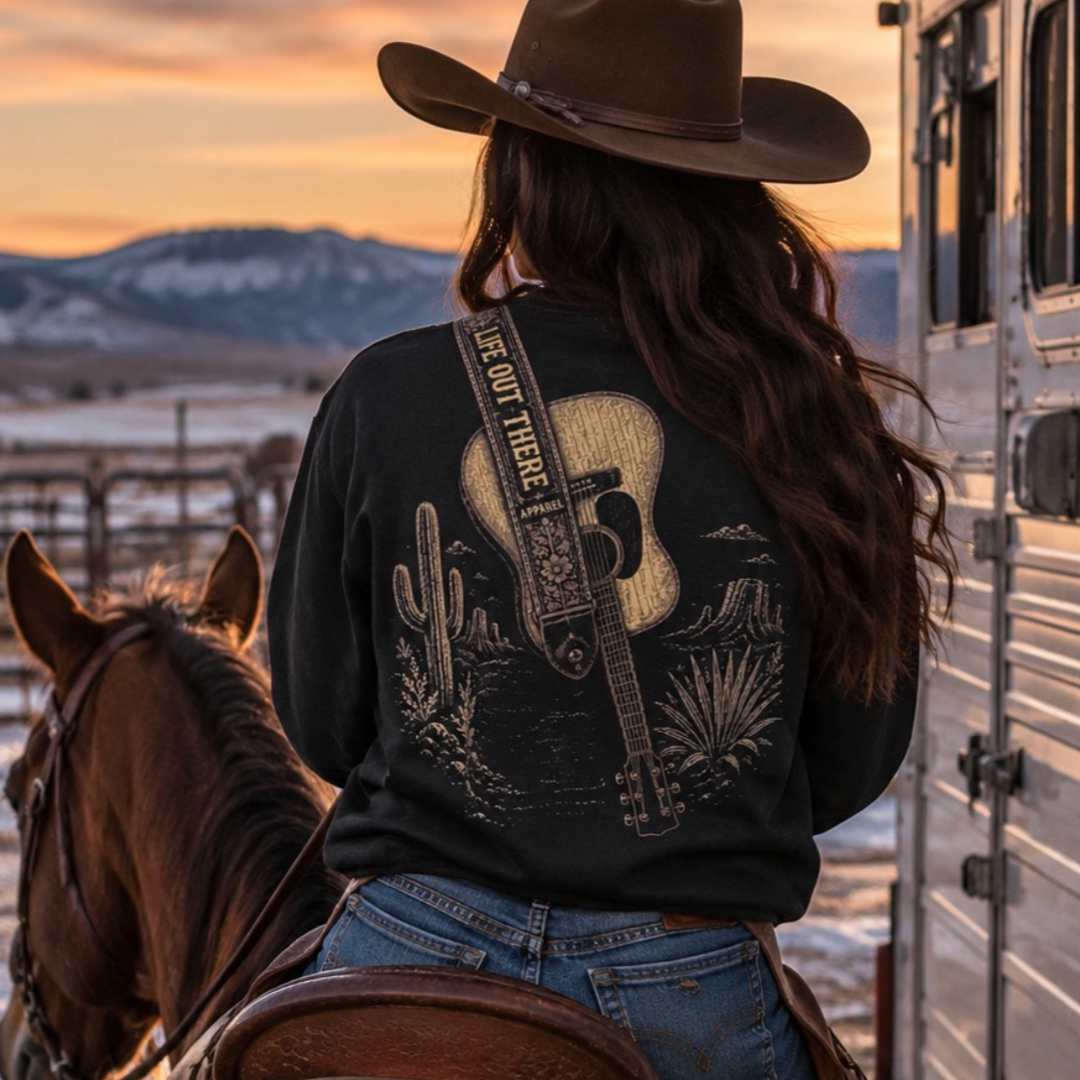 A person in a cowboy hat rides a horse at sunset, wearing the Life Out There Apparel Category Hurricane Crewneck Sweatshirt - Black, with mountains, a trailer, and pure country vibes in the background.