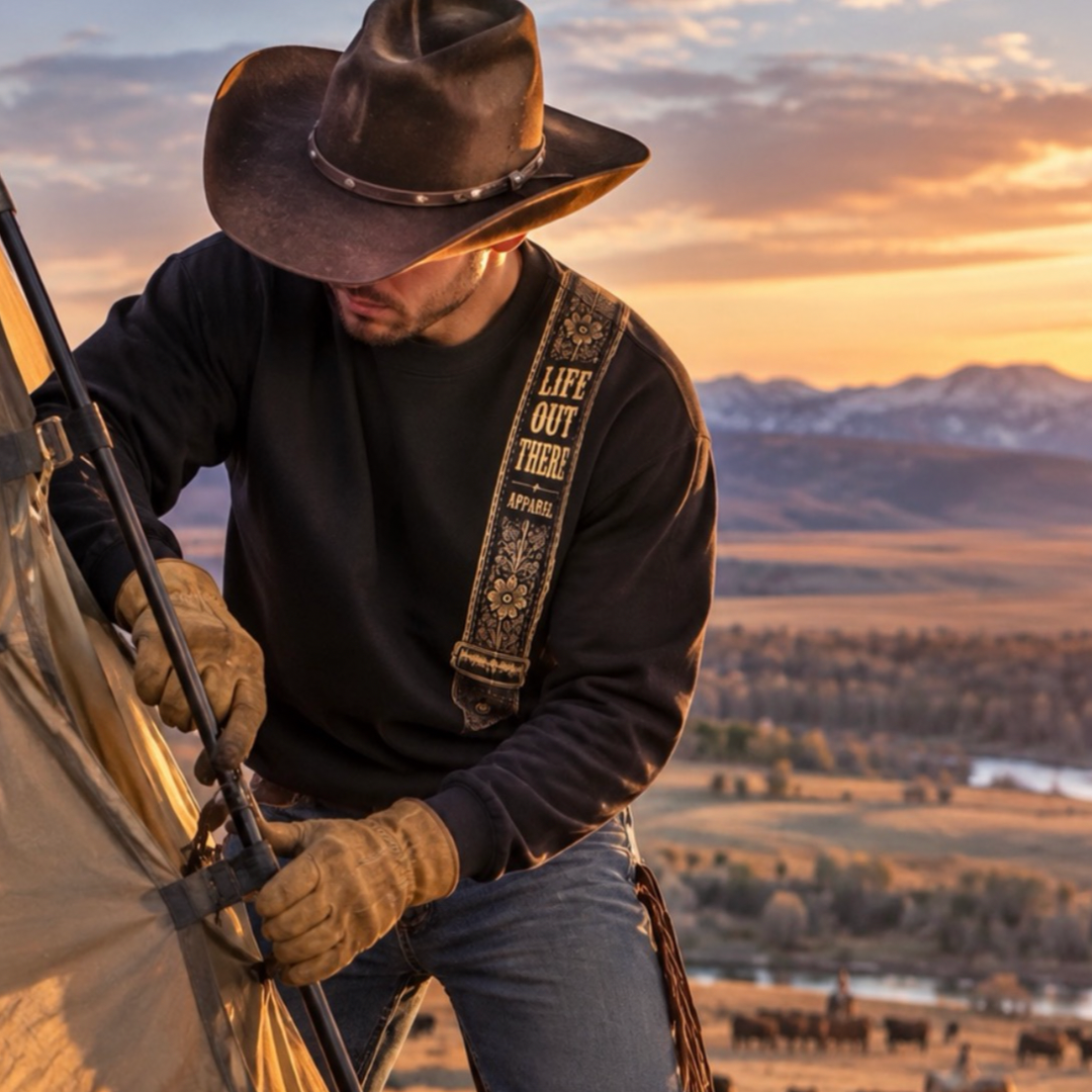 A cowboy in a brown hat and gloves sets up a tent at sunset, wearing the Category Hurricane Crewneck Sweatshirt - Black by Life Out There Apparel. Cattle graze nearby, adding to the classic country scene.