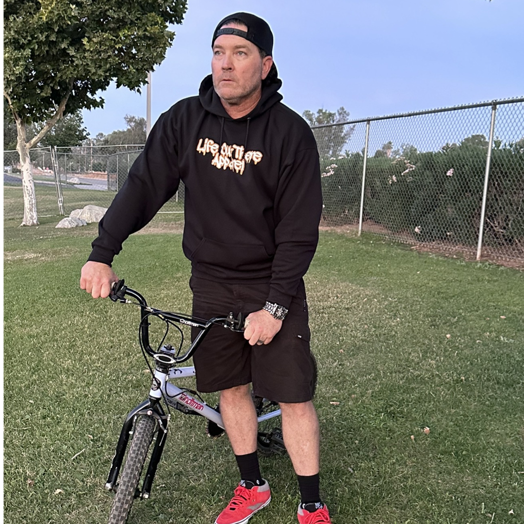 Life Out There Apparel “Fun is Not a Crime” Black Unisex hoodie has a skateboarder and bmx rider having fun on a half pipe skateboard ramp. A man  showing the front of the hoodie standing next to a bmx bike. 
