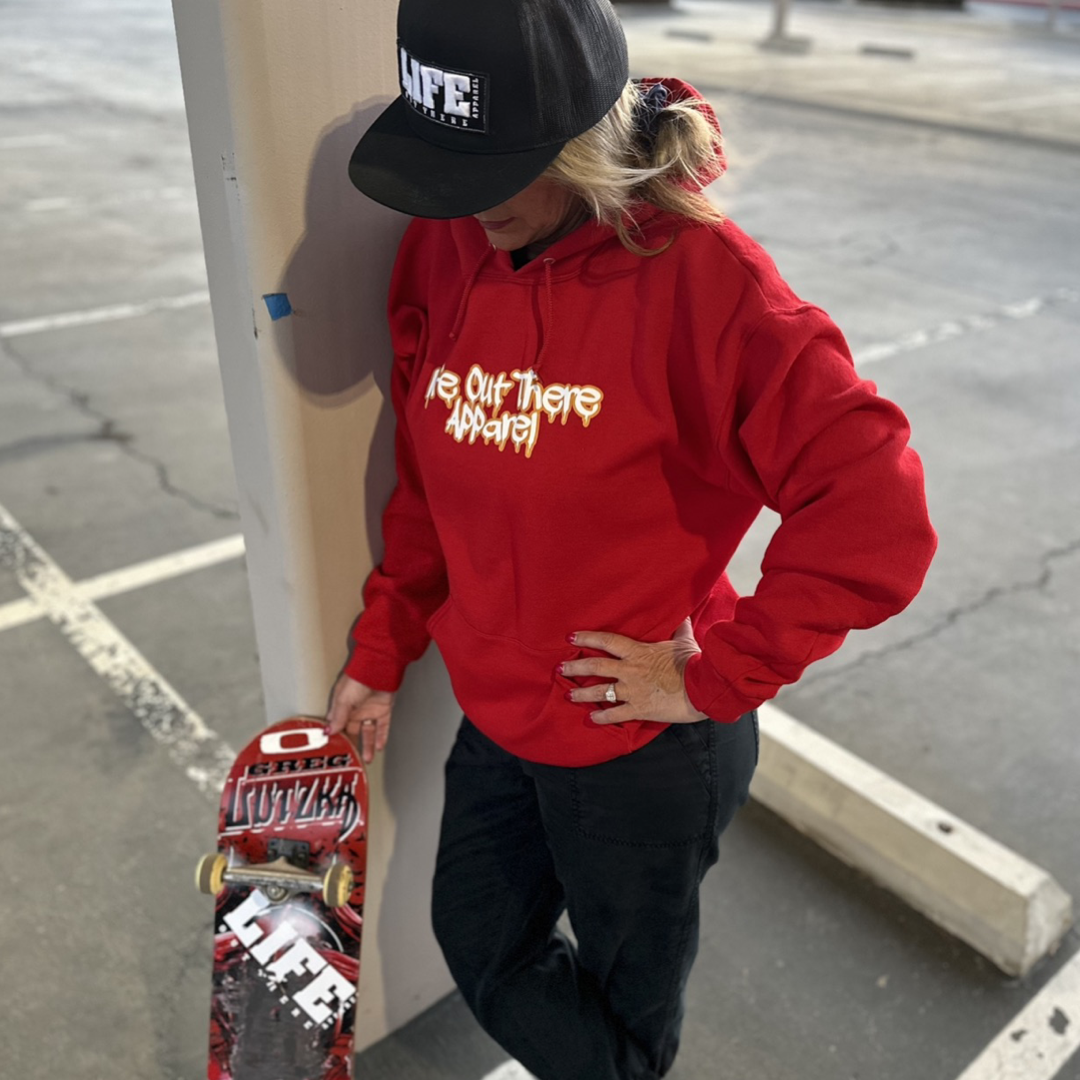 Life Out There Apparel “Fun is Not a Crime” Red Unisex hoodie has a skateboarder and bmx rider having fun on a half pipe skateboard ramp. A woman showing the front of the hoodie, holding a skateboard leaning up against a pillar. The front design looks like it’s spray painted. 