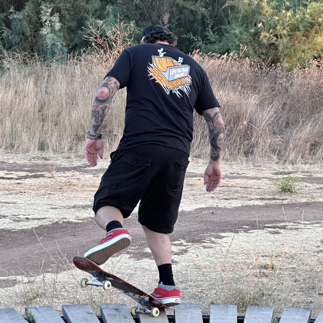 Life Out There Apparel “Fun is Not a Crime” Black Unisex tee shirt has a skateboarder and bmx rider having fun on a half pipe skateboard ramp. This picture has a guy standing in a nose blunt stance on a skateboard. 