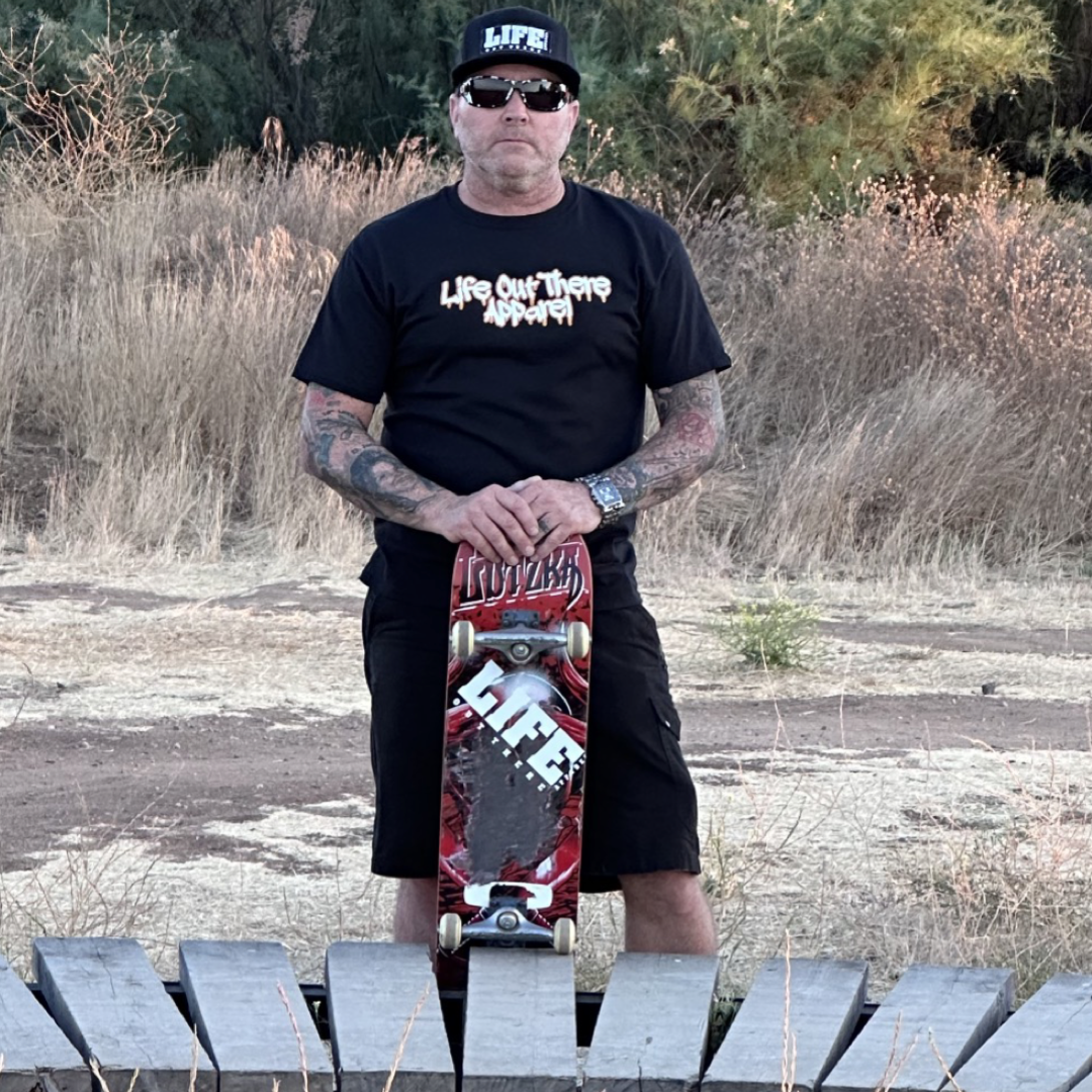 Life Out There Apparel “Fun is Not a Crime” Black Unisex tee shirt has a skateboarder and bmx rider having fun on a half pipe skateboard ramp. This picture has a guy standing holding a skateboard. 