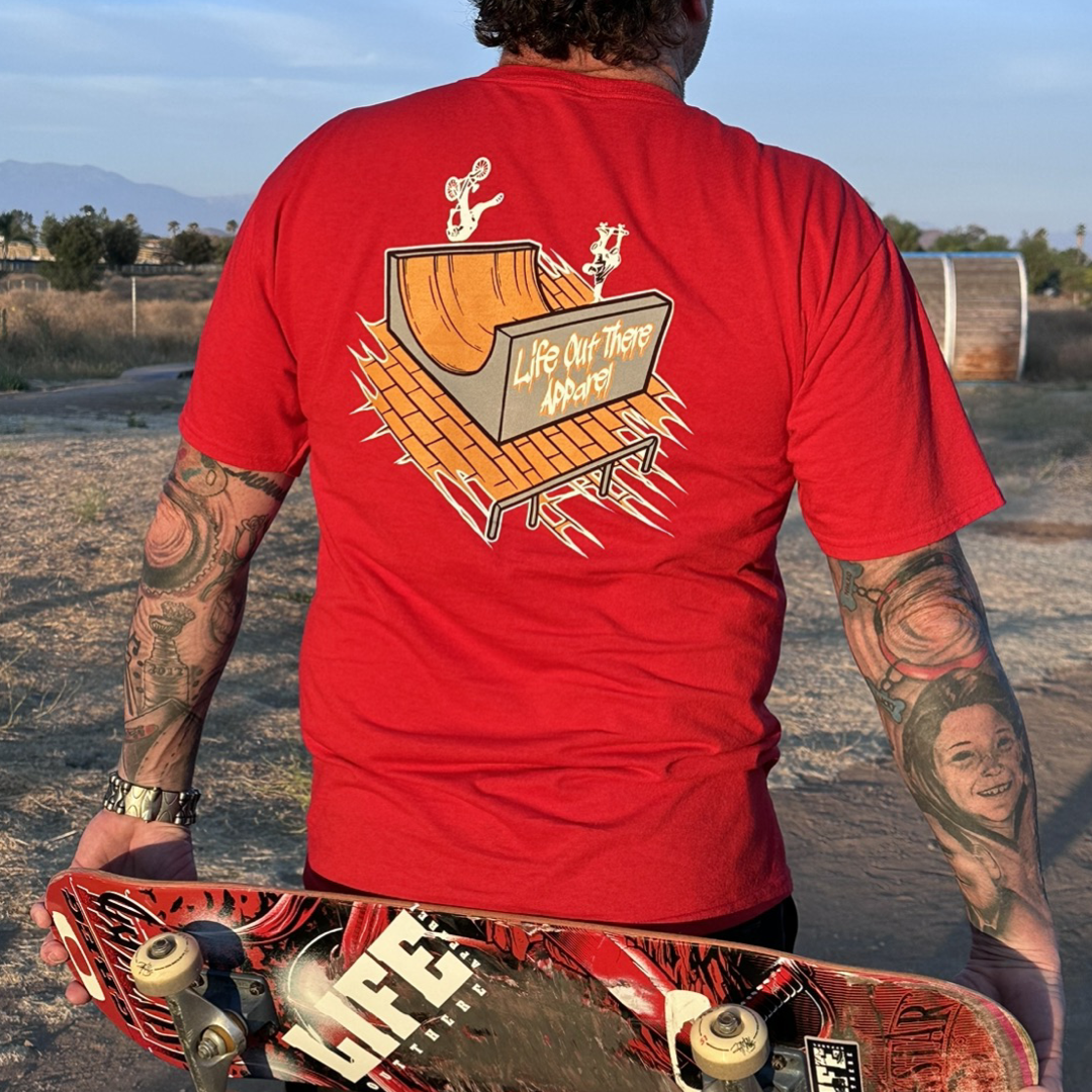 Life Out There Apparel “Fun is Not a Crime” Red Unisex tee shirt has a skateboarder and bmx rider having fun on a half pipe skateboard ramp. This picture has a man standing with a skateboard behind his back, at a park. 