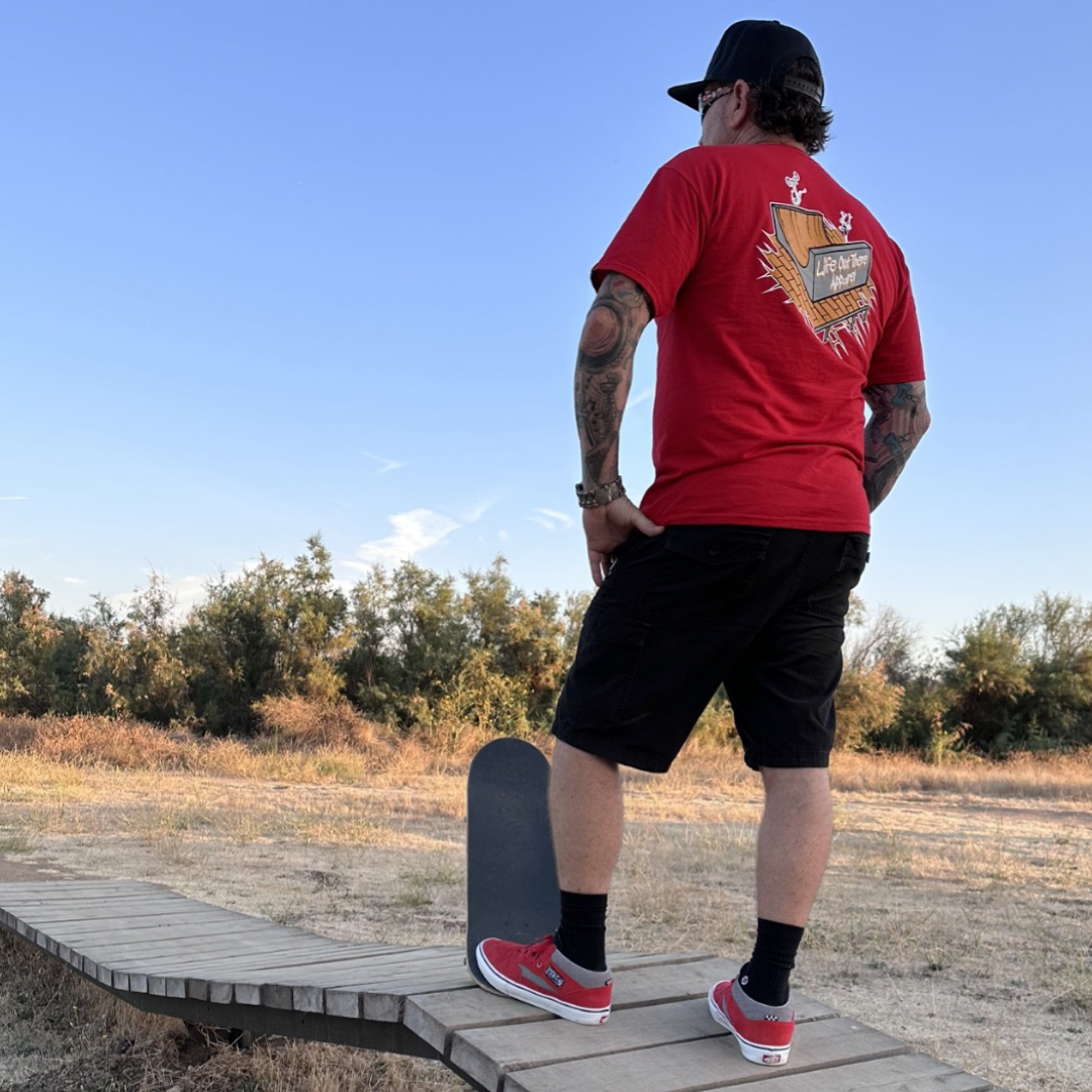 Life Out There Apparel “Fun is Not a Crime” Red Unisex tee shirt has a skateboarder and bmx rider having fun on a half pipe skateboard ramp. This picture has a man standing with a skateboard at a park. 