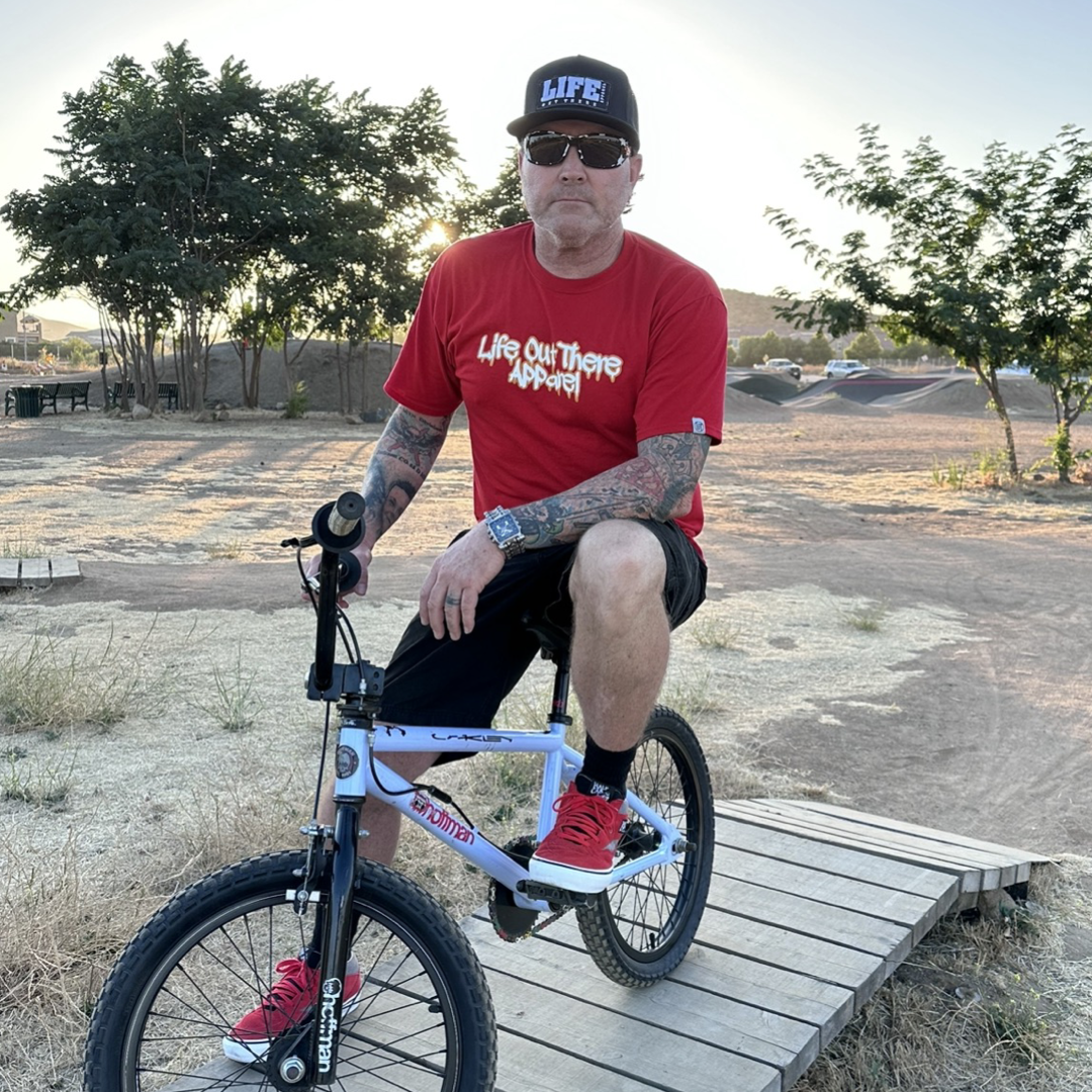 Life Out There Apparel “Fun is Not a Crime” Red Unisex tee shirt has a skateboarder and bmx rider having fun on a half pipe skateboard ramp. This picture has a man sitting on a bmx bike showing the front of the shirt. 