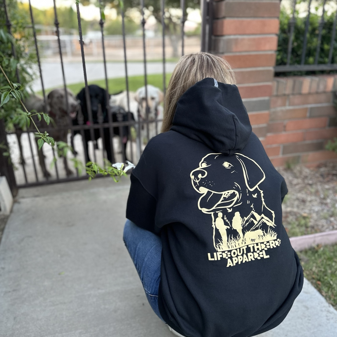Life Out There Apparel “Best Friend” Unisex hoodie in black. This is a women wearing the hoodie with all her dogs in front of her.  This is a tribute to our dogs, our best friend is a Labrador retriever dog on a hike with his humans. 