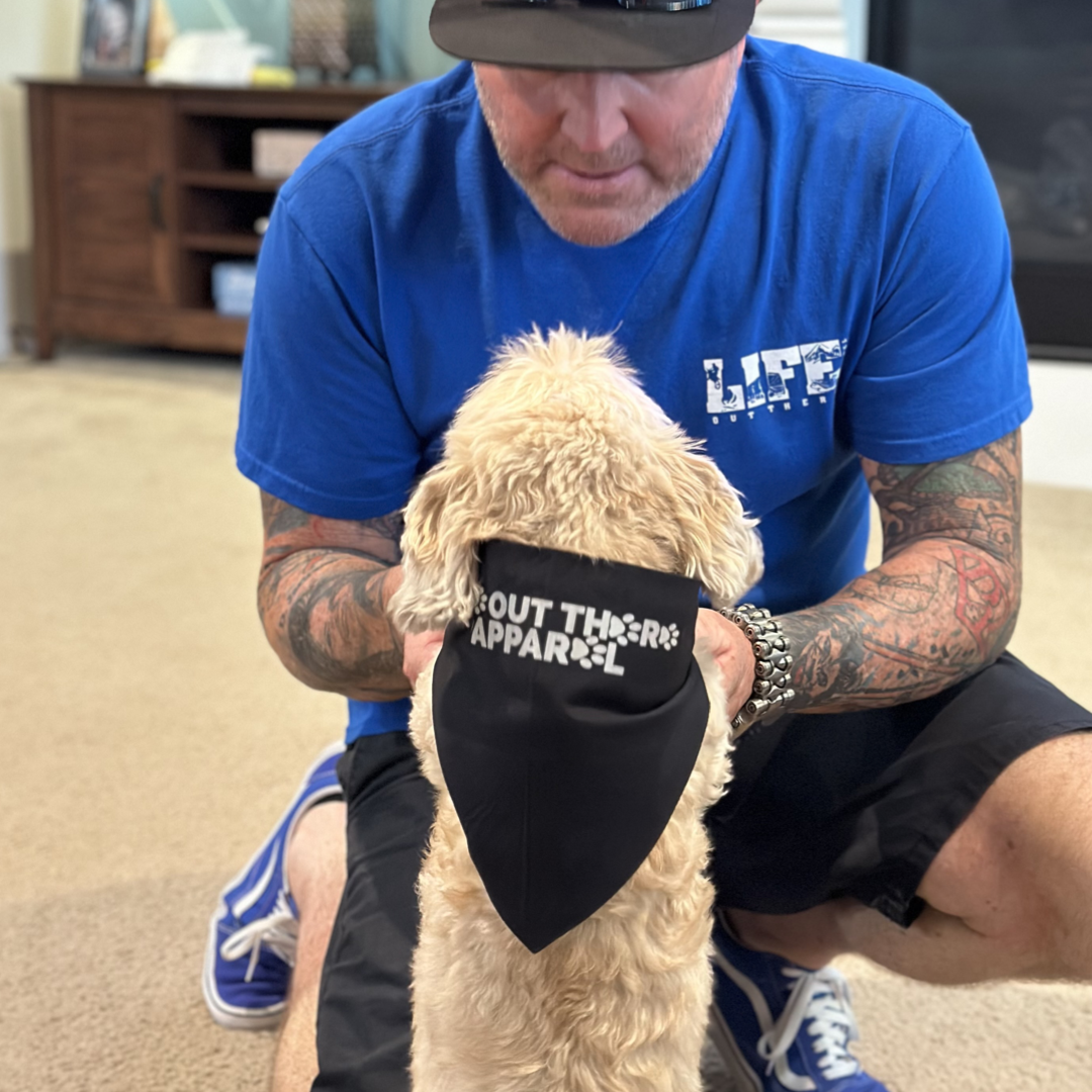 A man in a blue shirt and shorts kneels on the beige carpet, cradling his fluffy dog, his best friend. The dog is wearing the Best Friend Dog Bandanas - Black with Silver "REFLECTIVE" Print from Life Out There Apparel. In the background, a wooden cabinet adds warmth to the cozy room scene.