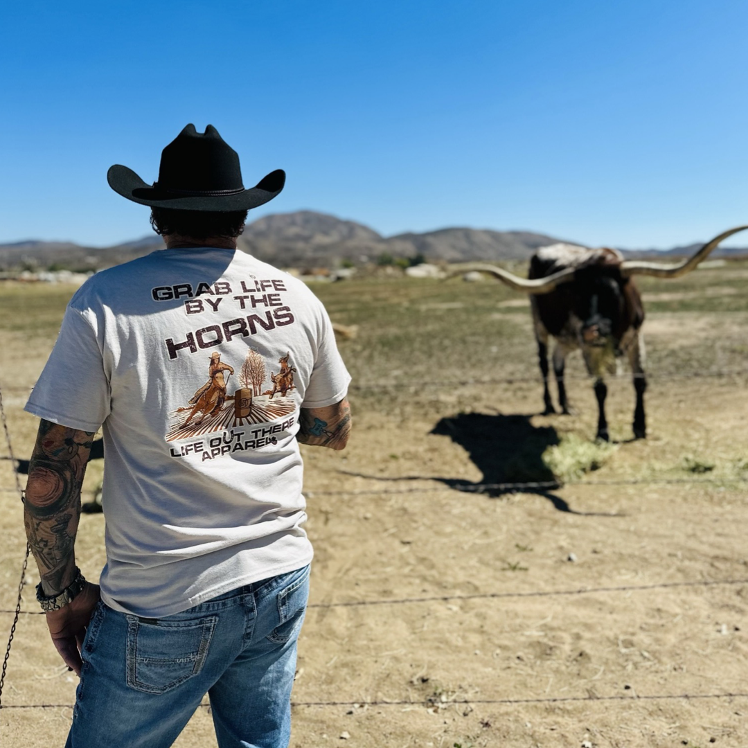 Life Out There Apparel Sandstone colored “Grab Life By The Horns” Unisex tee shirt has a girl barrel racer getting ready to turn around a barrel with a guy riding a bull behind her.  This shows a guy wearing a cowboy hat, standing in front of a group of long horn bulls, showing the back of the tee. 