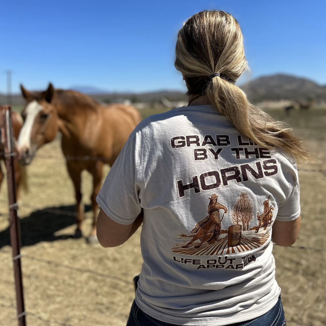 Life Out There Apparel Sandstone colored “Grab Life By The Horns” Unisex tee shirt has a girl barrel racer getting ready to turn around a barrel with a guy riding a bull behind her.  This shows a women showing the back of the tee standing in front of some horses. 