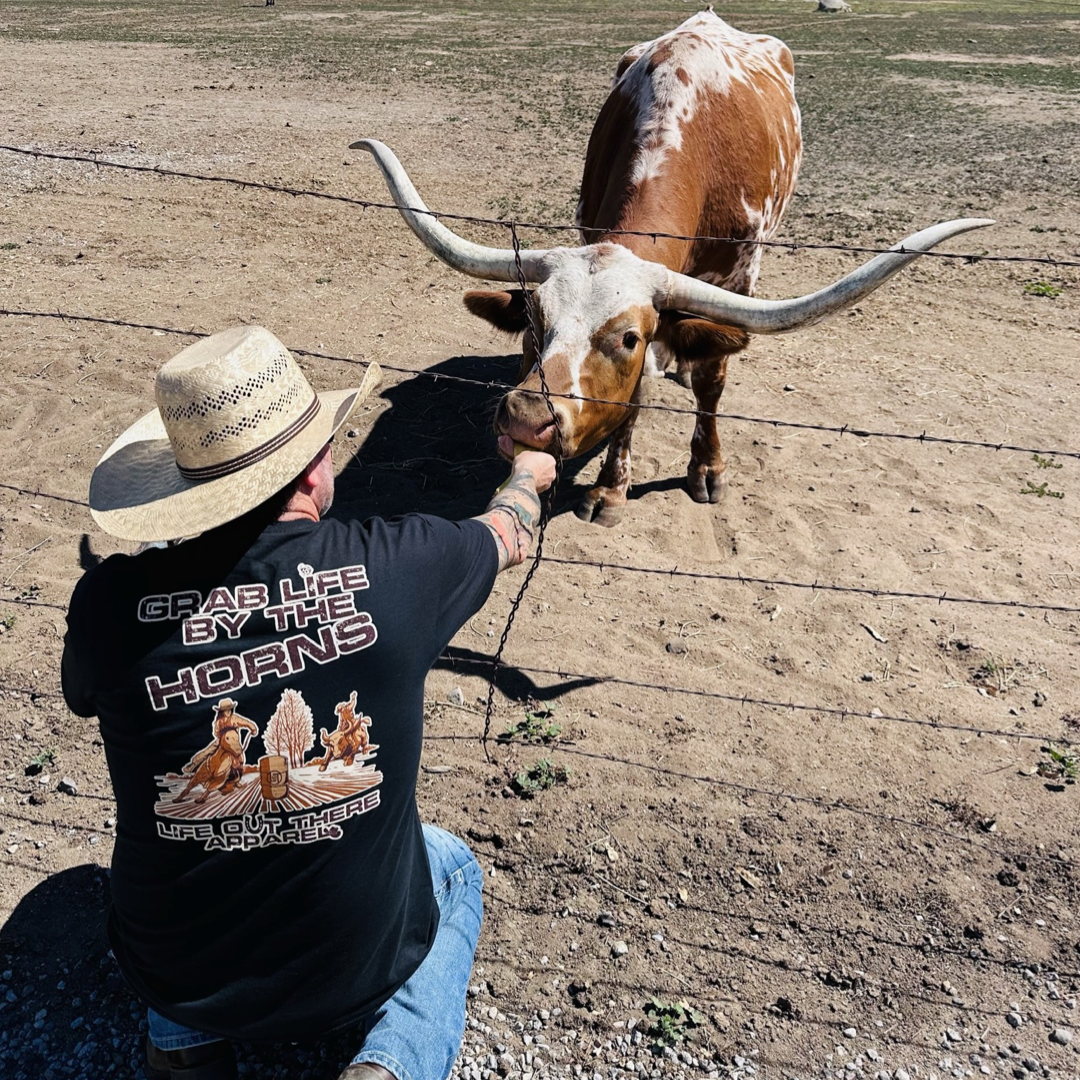 Life Out There Apparel “Grab Life By The Horns” Black Unisex tee shirt has a girl barrel racer getting ready to turn around a barrel with a guy riding a bull behind her.  This shows a cowboy wearing the shirt feeding a banana to a long horn bull. 