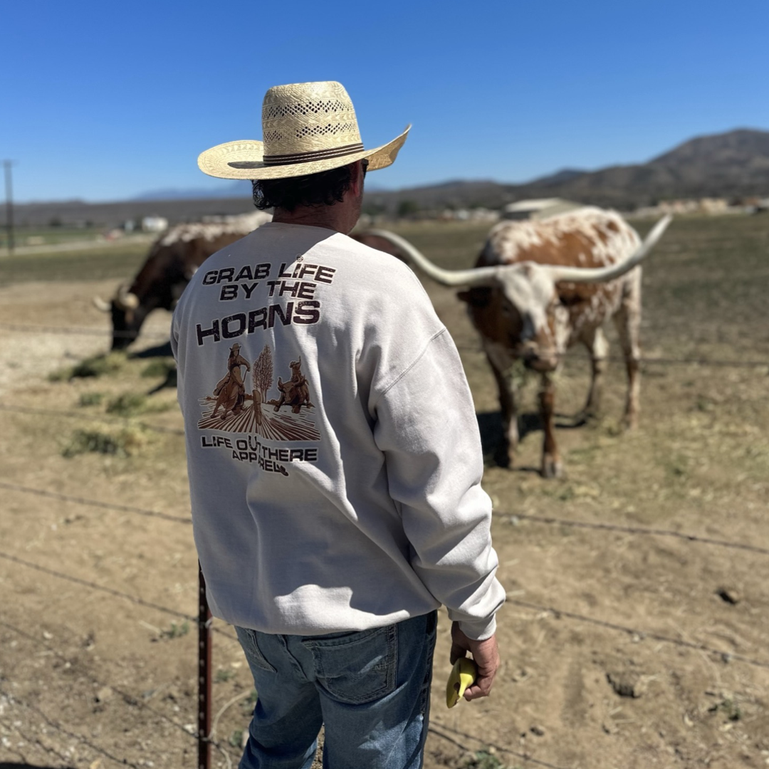 Life Out There Apparel Sandstone colored “Grab Life By The Horns” Unisex Crew Neck Sweatshirt has a girl barrel racer getting ready to turn around a barrel with a guy riding a bull behind her.  This shows a guy wearing the sweatshirt standing in front of a bull. 
