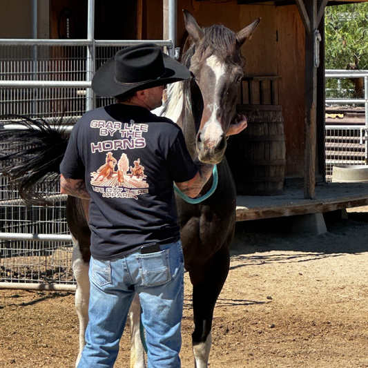 Life Out There Apparel “Grab Life By The Horns” Black Unisex tee shirt has a girl barrel racer getting ready to turn around a barrel with a guy riding a bull behind her.  This shows a cowboy wearing the tee shirt, showing the back, standing with his horse. 