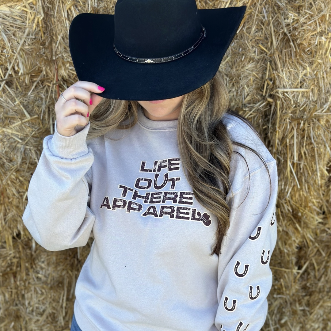 Life Out There Apparel Sandstone colored “Grab Life By The Horns” Unisex Crew Neck Sweatshirt has a girl barrel racer getting ready to turn around a barrel with a guy riding a bull behind her.  This shows a girl wearing the sweatshirt with a cowboy hat on in front of a stack of hay.   
