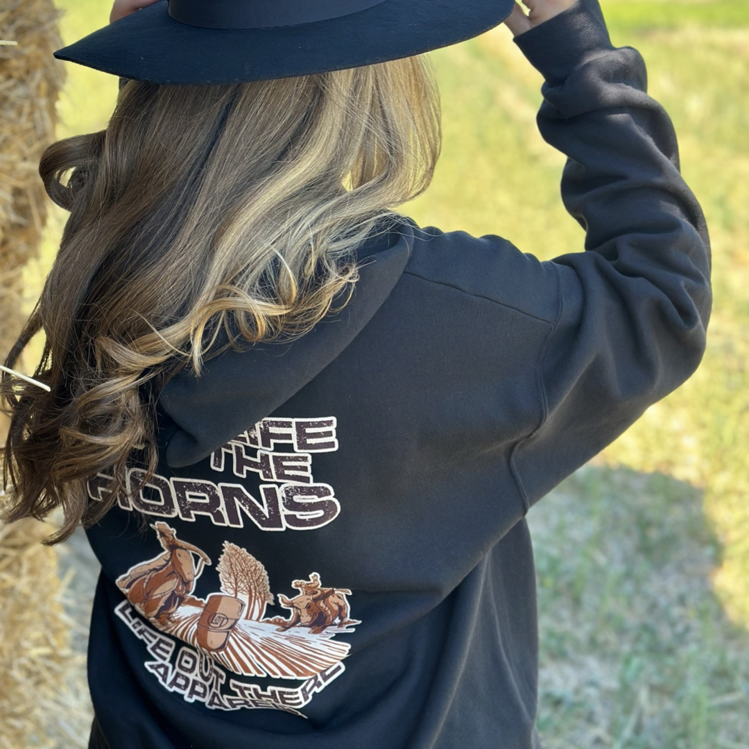 Life Out There Apparel “Grab Life By The Horns” Black Unisex Hoodie has a girl barrel racer getting ready to turn around a barrel with a guy riding a bull behind her.  This shows a girl wearing the black hoodie with a cowboy hat on leaning against a stack of hay. 