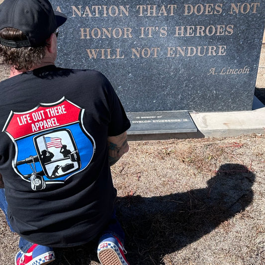 Life Out There Apparel “Leave It Behind” Black Unisex tee shirt has a truck side mirror with the reflection of a male, female & K9 soldiers saluting the American flag - bringing awareness to P. T. S. D. (post traumatic stress disorder). This is a photo of a man kneeling at a memorial plaque at an air museum. 