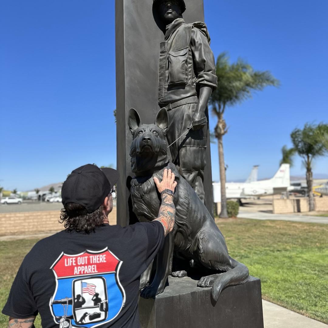 Life Out There Apparel “Leave It Behind” Black Unisex tee shirt has a truck side mirror with the reflection of a male, female & K9 soldiers saluting the American flag - bringing awareness to P. T. S. D. (post traumatic stress disorder). This is a photo of a man at a K9 memorial statue paying tribute. 