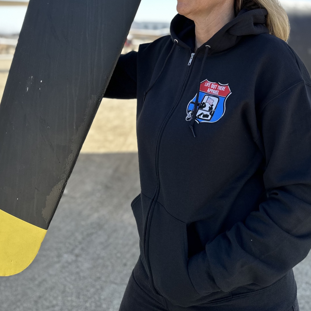 A Black Unisex “Zip Up” hoodie from Life Out There Apparel - “Leave It Behind”  has a truck side mirror with the reflection of a male, female & K9 soldiers saluting the American flag - bringing awareness to P. T. S. D. (post traumatic stress disorder). This is a photo of a woman wearing the hoodie showing the front design, standing next to a military airplane learning on the propeller.  