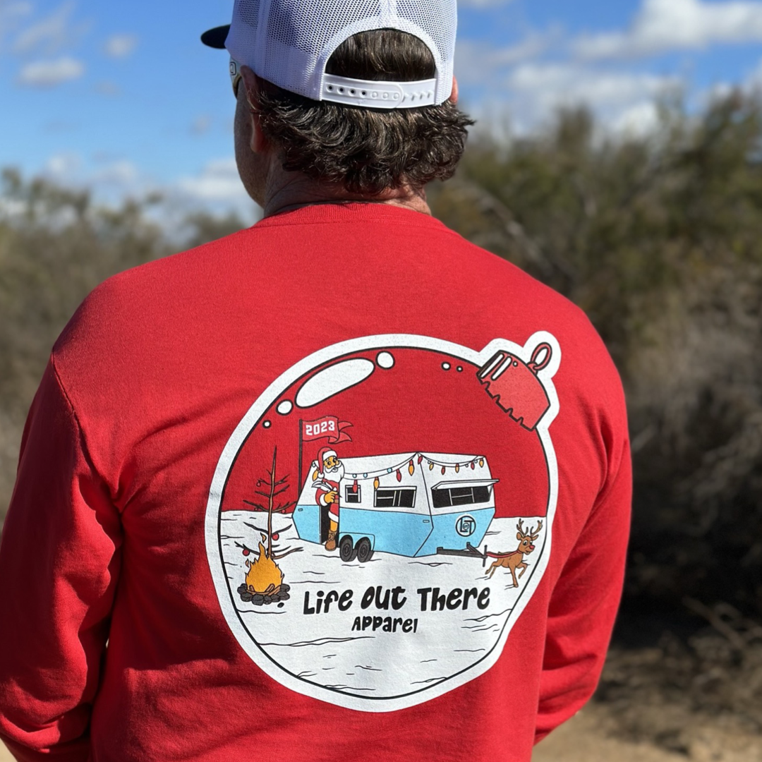 Life Out There Apparel “December 26th” red Unisex long sleeve tee shows Santa Clause camping in the desert, in a travel trailer, while relaxing after the big day and burning his Christmas tree, all inside a Christmas ornament. A man showing the back of the shirt.  