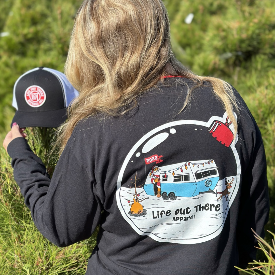 Life Out There Apparel “December 26th” black Unisex long sleeve tee shows Santa Clause camping in the desert, in a travel trailer, while relaxing after the big day and burning his Christmas tree, all inside a Christmas ornament. A women showing the back of the shirt & she’s holding a trucker hat. 