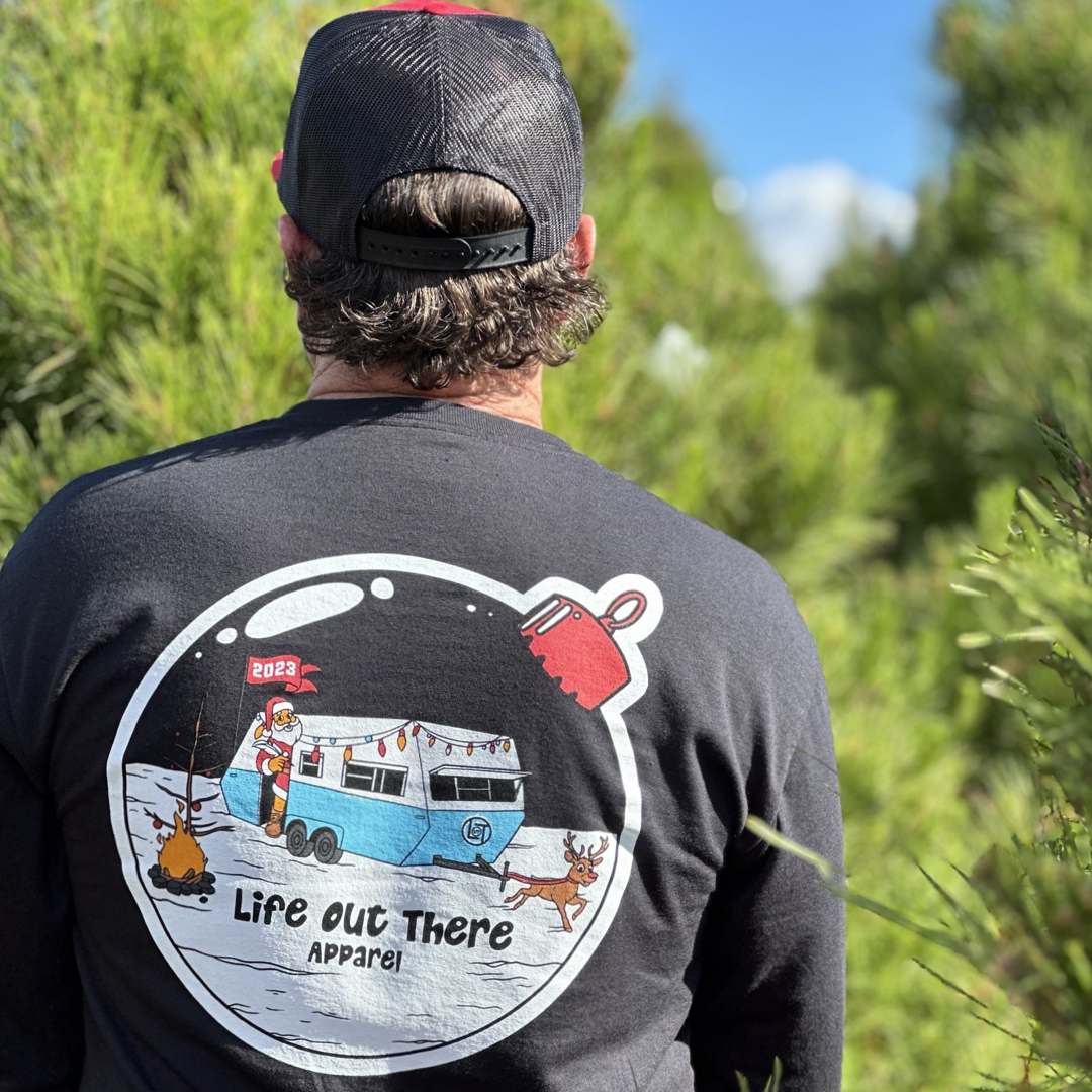 Life Out There Apparel “December 26th” black Unisex long sleeve tee shows Santa Clause camping in the desert, in a travel trailer, while relaxing after the big day and burning his Christmas tree, all inside a Christmas ornament. A man showing the back of the shirt in front of a Christmas tree. 
