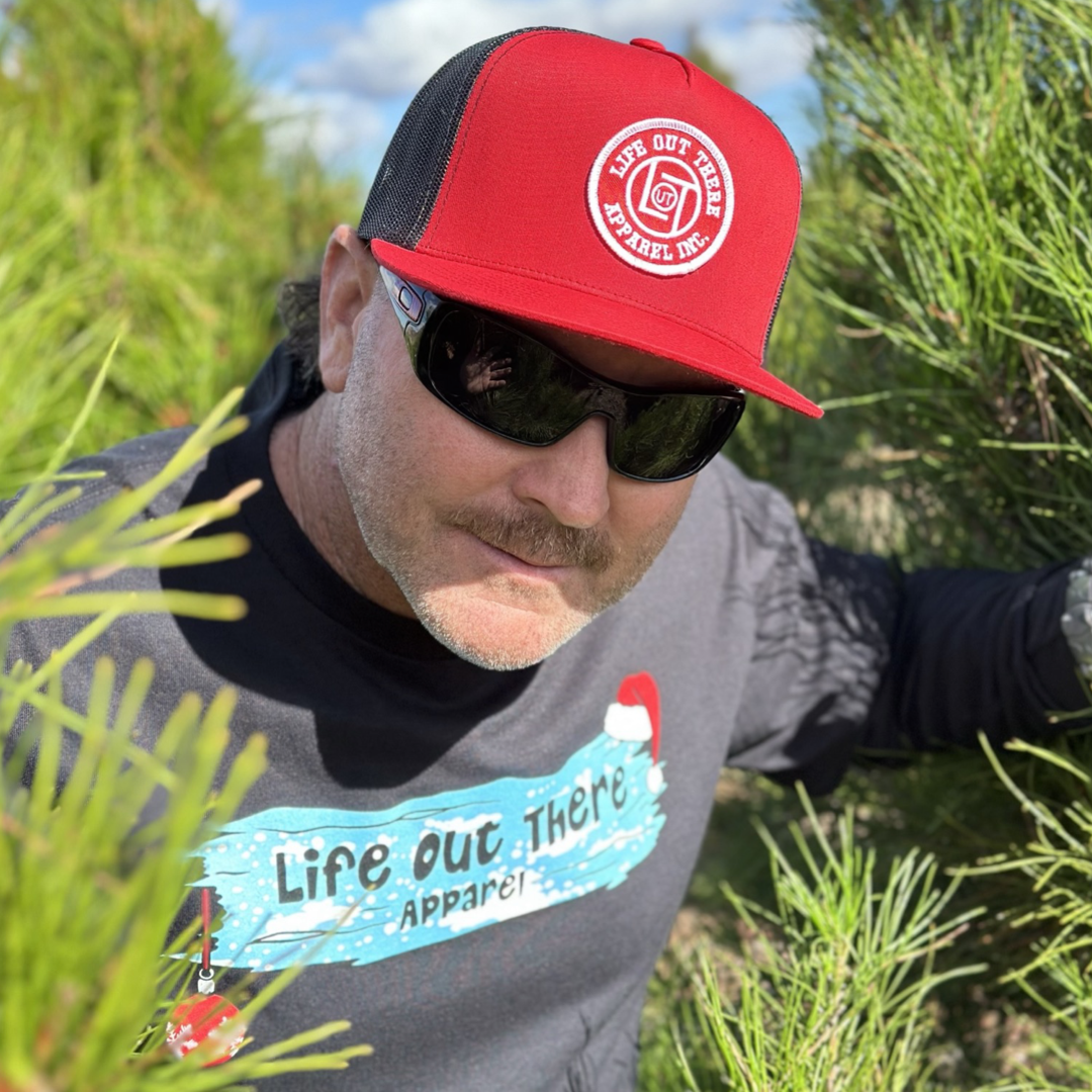 Life Out There Apparel “December 26th” black Unisex long sleeve tee shows Santa Clause camping in the desert, in a travel trailer, while relaxing after the big day and burning his Christmas tree, all inside a Christmas ornament. A man showing the front of the shirt peeking through a Christmas tree.