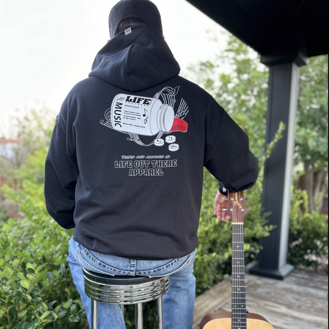 Life Out There Apparel “Music Is Medicine “ The Black Unisex Pullover Hoodie design is a medicine pill bottle tipped over with pills that have fallen out, that have music notes on them. This photo has a man in a chair  leaning against a guitar showing the back of the hoodie.