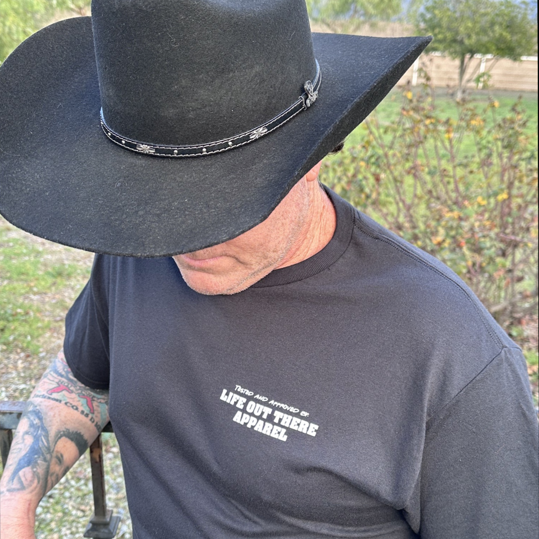 Life Out There Apparel “Music Is Medicine “ Black Unisex  tee shirt is a medicine pill bottle tipped over with pills that have fallen out, that have music notes on them. This photo has a man wearing a cowboy hat showing the front of the shirt on a ranch. 