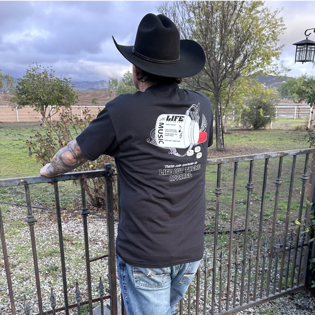 Life Out There Apparel “Music Is Medicine “ Black Unisex  tee shirt is a medicine pill bottle tipped over with pills that have fallen out, that have music notes on them. This photo has a man wearing a cowboy hat showing the back of the shirt, looking out onto a ranch. 