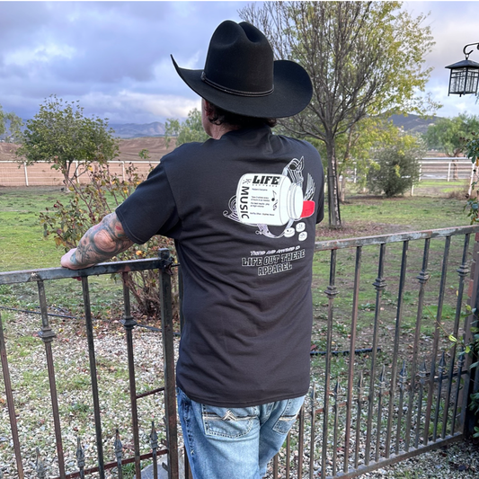 Wearing the Life Out There Apparel "Music Is Medicine" T Shirt in black, a person in a cowboy hat stands by a metal fence, looking at a grassy yard with trees and mountains. The shirt features guitar art and inspiring music-themed text.