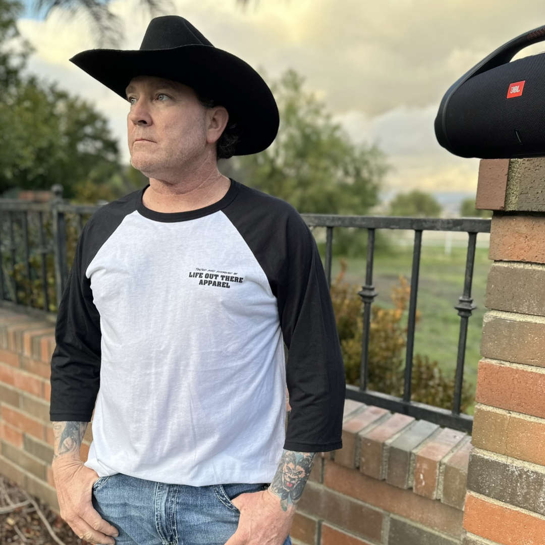 Black & White Life Out There Apparel “Music Is Medicine “ Unisex Raglan 3/4 tee shirt is a medicine pill bottle tipped over with pills that have fallen out, that have music notes on them. This photo has a man showing the front of the shirt standing in front of a fence.  