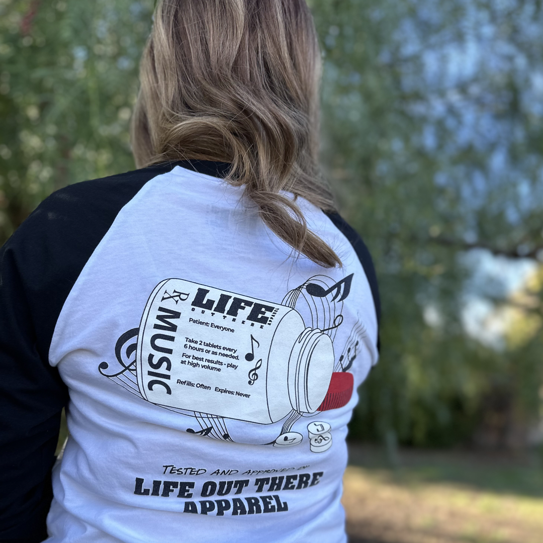 Black & White Life Out There Apparel “Music Is Medicine “ Unisex Raglan 3/4 tee shirt is a medicine pill bottle tipped over with pills that have fallen out, that have music notes on them. This photo has a woman showing the back of the shirt standing in a field.   
