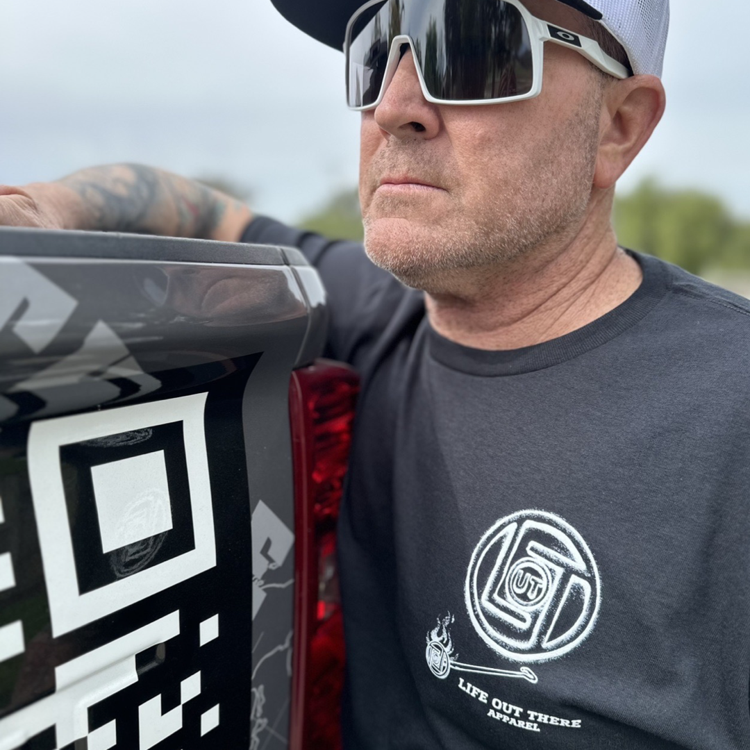 Life Out There Apparel “Branded” Unisex black tee shirt is a round logo of the companies initials, made with a hot branding iron. This is a photo of a man wearing the shirt leaning up, against his truck. 
