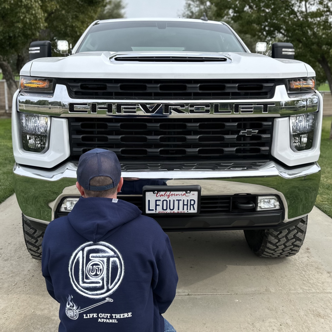 Life Out There Apparel “Branded” Unisex navy colored hoodie is a round logo of the companies initials, made with a hot branding iron. This is a photo of a guy wearing the hoodie squatting in front of a truck. 