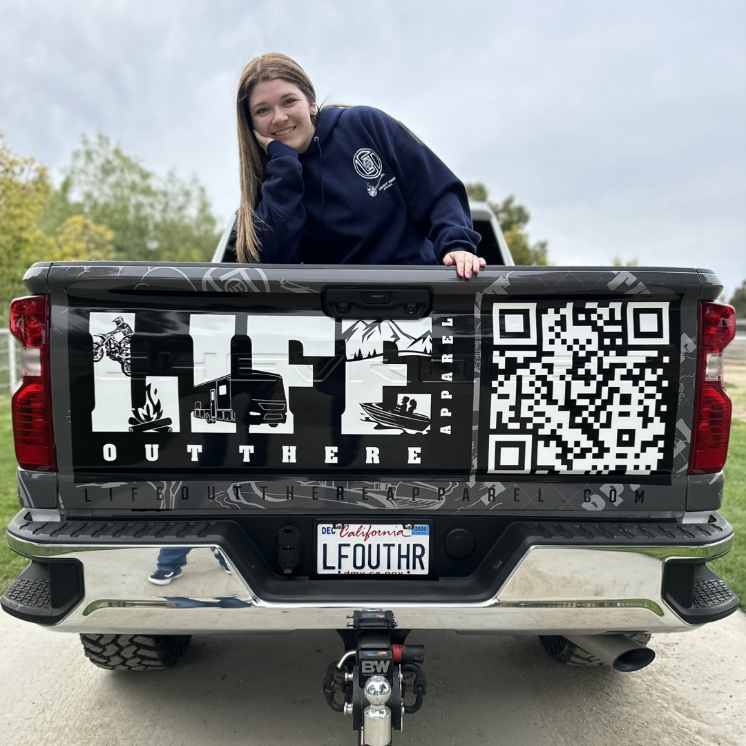 Life Out There Apparel “Branded” Unisex navy blue hoodie is a round logo of the companies initials, made with a hot branding iron. This is a young woman wearing the hoodie sitting in the bed of a truck.   