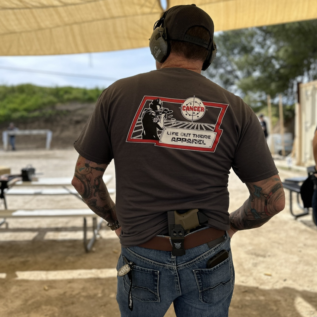 Life Out There Apparel “Kill Cancer” Charcoal Grey Unisex tee shirt has picture of a man shooting a sign that says cancer on it & the bullet rounds coming out of the gun have  “chemo” written on them. This picture is a man at the gun range waiting his turn to shoot, showing the back of the shirt. 