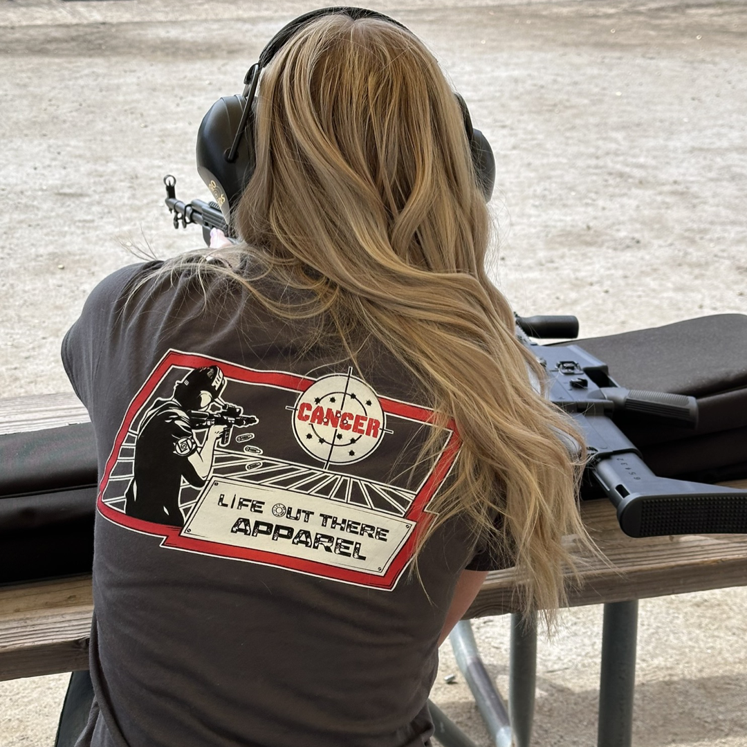 Life Out There Apparel “Kill Cancer” Charcoal Grey Unisex tee shirt has picture of a man shooting a sign that says cancer on it & the bullet rounds coming out of the gun have  “chemo” written on them. This picture is a woman at the gun range waiting her turn to shoot at the range, showing the back of the shirt. 