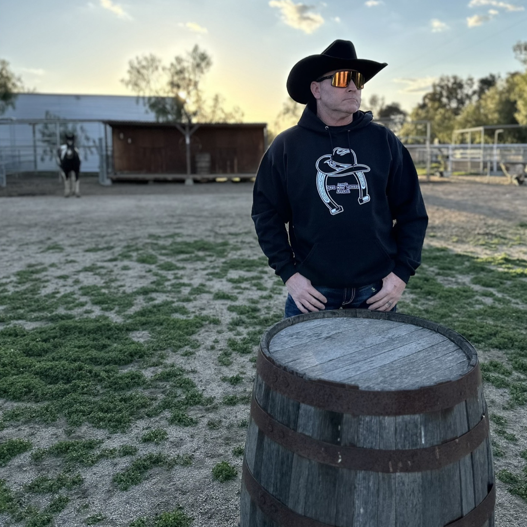 Black Unisex hoodie from Life Out There Apparel “ONWARD & UPWARD” Tour - this design is a country concert design with a cowboy hat & horseshoe on the front and back with every Life Out There Apparel launch for the past 2 years. This photo shows a man wearing a cowboy hat looking out into a ranch with a horse in the background.  