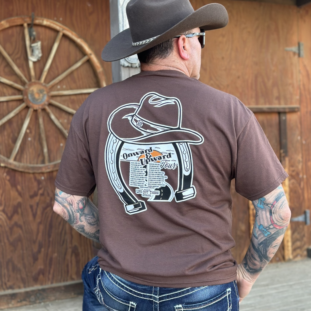 “ONWARD & UPWARD” Tour :
The Chocolate Brown Life Out There Apparel “ONWARD & UPWARD” Tour Unisex tee is a country concert design with a cowboy hat & horseshoe on the front and back, with all the design names from the past 2 years (instead of city & tours dates) This photo is of a man wearing a cowboy hat standing in front of a tack / feed room on a ranch. 