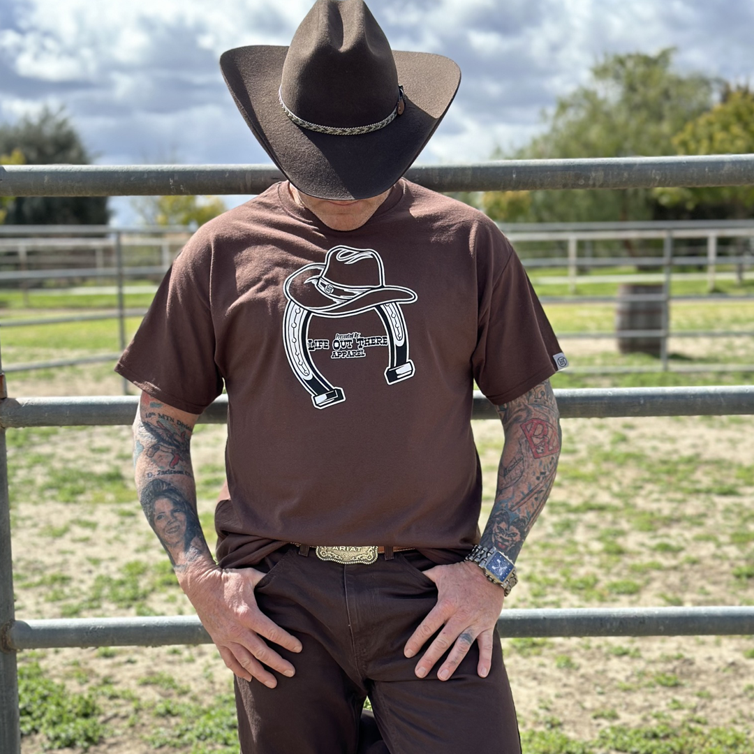 “ONWARD & UPWARD” Tour :
The Life Out There Apparel “ONWARD & UPWARD” Tour Unisex tee in Chocolate Brown is a country concert design with a cowboy hat & horseshoe on the front and back, with all the design names from the past 2 years (instead of city & tours dates) This photo is of a man wearing a cowboy hat standing on a ranch next to a round pin.  