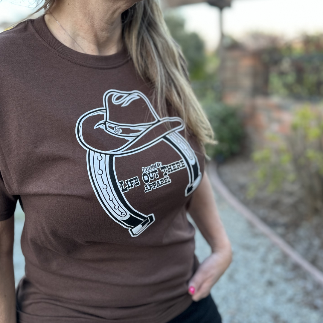 “ONWARD & UPWARD” Tour :
The Chocolate Brown Life Out There Apparel “ONWARD & UPWARD” Tour Unisex tee in is a country concert design with a cowboy hat & horseshoe on the front and back, with all the design names from the past 2 years (instead of city & tours dates) This photo is of a woman wearing a cowgirl standing on a ranch. 