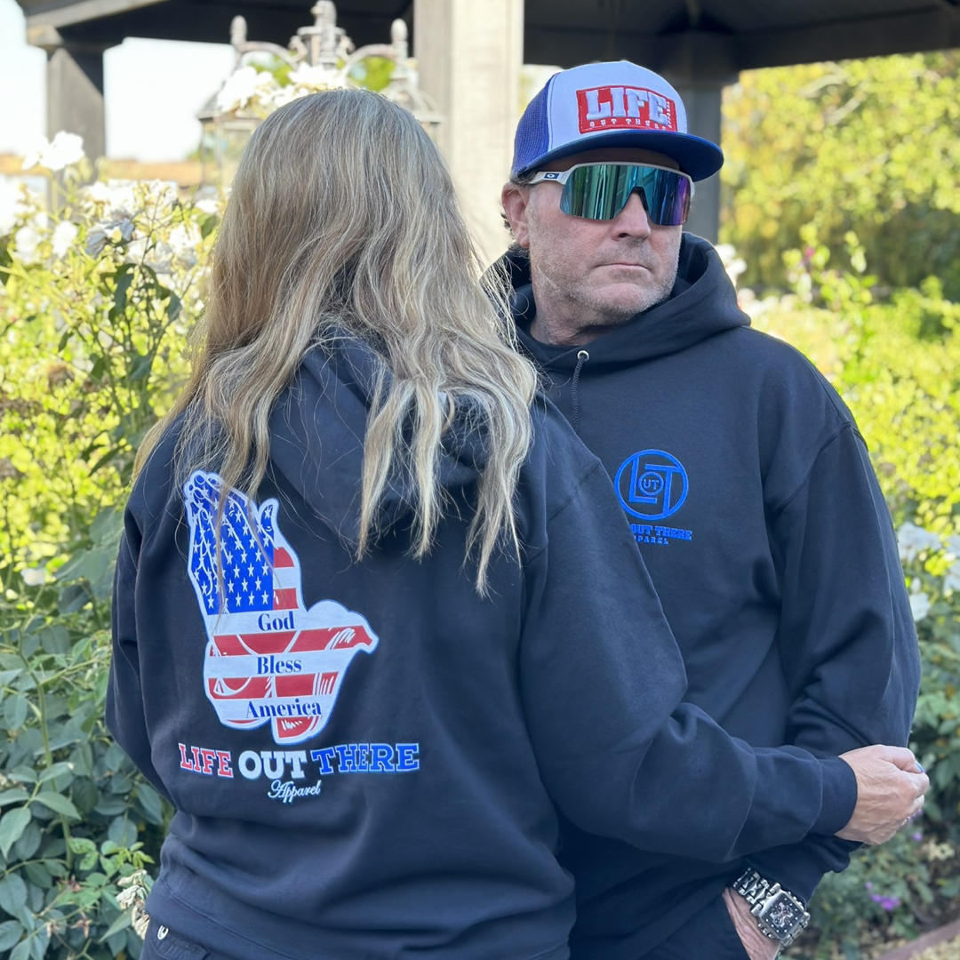 Two individuals wear black hoodies from Life Out There Apparel, ideal for 4th of July celebrations. One wears the "Unisex God Bless America Hoodie - Black," featuring the phrases "God Bless America" and "Life Out There," while the other showcases a logo on the front. The person to the left boasts long hair, while their companion is accessorized with sunglasses and a cap.