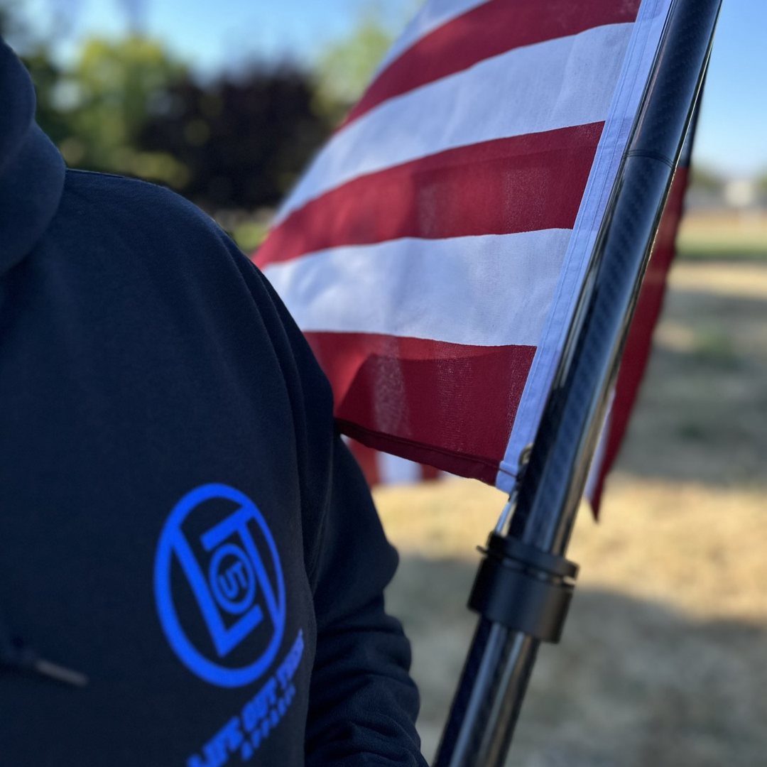 A person wearing the Unisex God Bless America Hoodie from Life Out There Apparel, featuring a blue circular logo, stands beside an American flag. The scene captures the spirit of the 4th of July, with the flag attached to a pole and set against a backdrop of blurred greenery and trees.