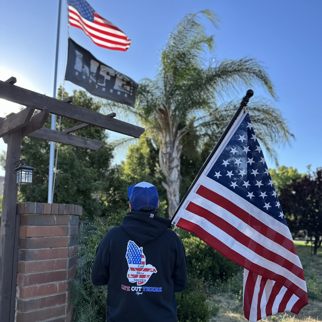 A person stands holding an American flag, dressed in a Unisex God Bless America Hoodie by Life Out There Apparel, featuring a USA outline design. They gaze toward another American flag and a black flag flying on a pole. Surrounded by palm trees under a clear blue sky, the scene beautifully captures the essence of God Bless America.