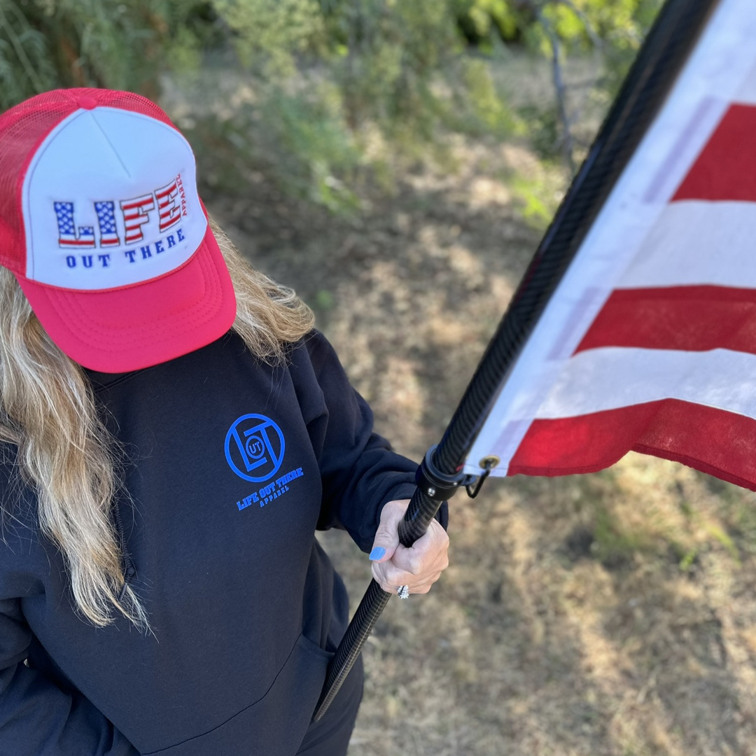 A person with long blonde hair wearing a Life Out There Apparel cap and a Unisex God Bless America Hoodie in black holds an American flag upright. The background of trees and grass sets a patriotic scene, echoing the spirit of the 4th of July in the United States of America.