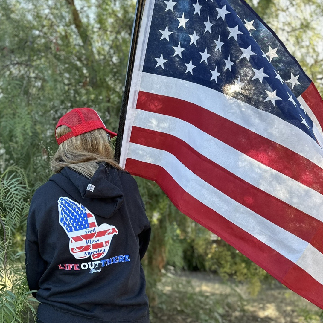 A person wearing the Life Out There Apparel's Unisex God Bless America Hoodie in black, featuring a USA-shaped design, holds a large American flag while facing away. The lush green trees in the background beautifully capture the spirit of the 4th of July and resonate with "God Bless America.