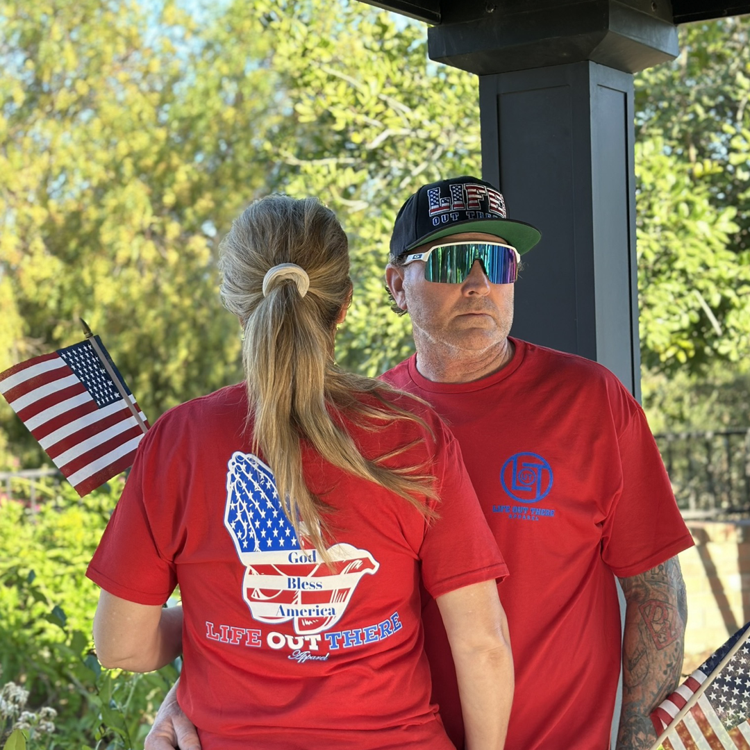 A man and woman stand under a shaded structure, both wearing Life Out There Apparel's Unisex God Bless America T Shirt in red, proudly holding American flags. The man sports a cap with sunglasses, while the woman wears her hair in a ponytail. Greenery and trees form the backdrop, celebrating the spirit of 4th of July 2024.