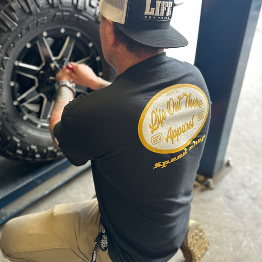 A person in a Life Out There Apparel Speed Shop T Shirt—Black kneels next to a large off-road tire, appearing to work on it in a workshop setting.