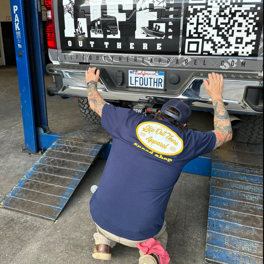 A man in a navy Speed Shop T Shirt by Life Out There Apparel kneels behind a truck in a garage, inspecting its rear bumper and California plate "LFOUTHR." The tailgate displays a large QR code and bold automotive apparel graphics.