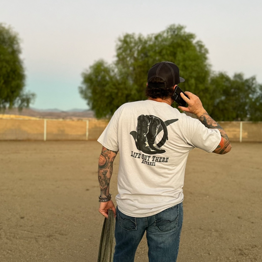 A tattooed man wearing a silver Boot Check T Shirt from Life Out There Apparel and a cap stands outdoors on dirt, holding something in his left hand and talking on the phone. Trees and a fence can be seen in the background.