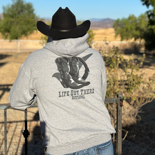 A person wearing a Life Out There Apparel Unisex Boot Check Hoodie in Oxford Grey and a black cowboy hat stands by a fence. The backdrop features trees and dry, rolling hills under a clear blue sky, subtly nodding to the spirit of prevention month through fashion.